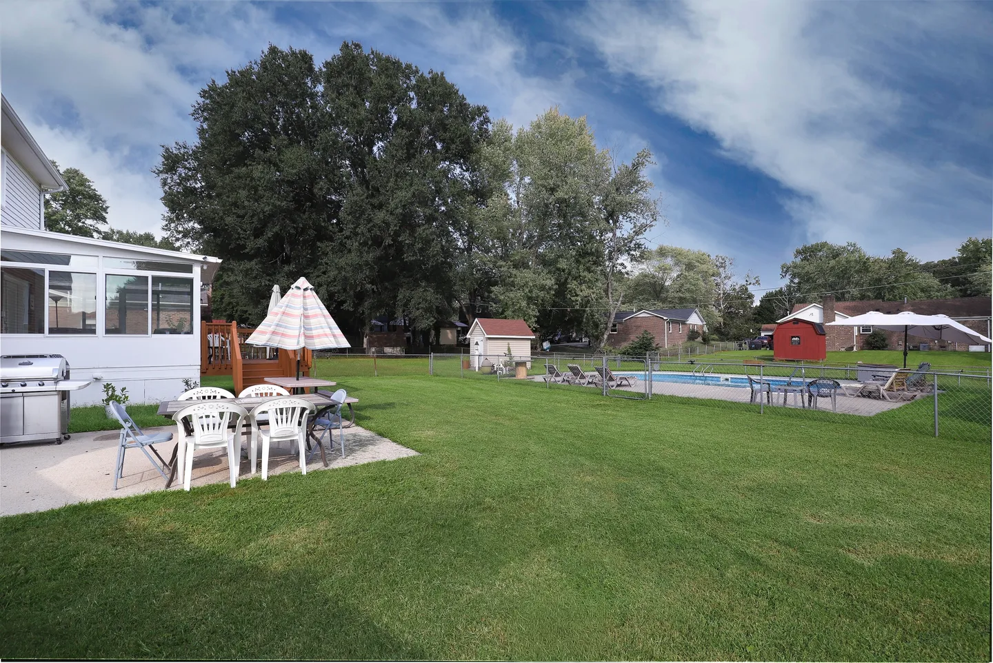 Spacious grassy backyard with a concrete patio featuring outdoor dining furniture, a grill, and an enclosed fenced area with an in-ground swimming pool.