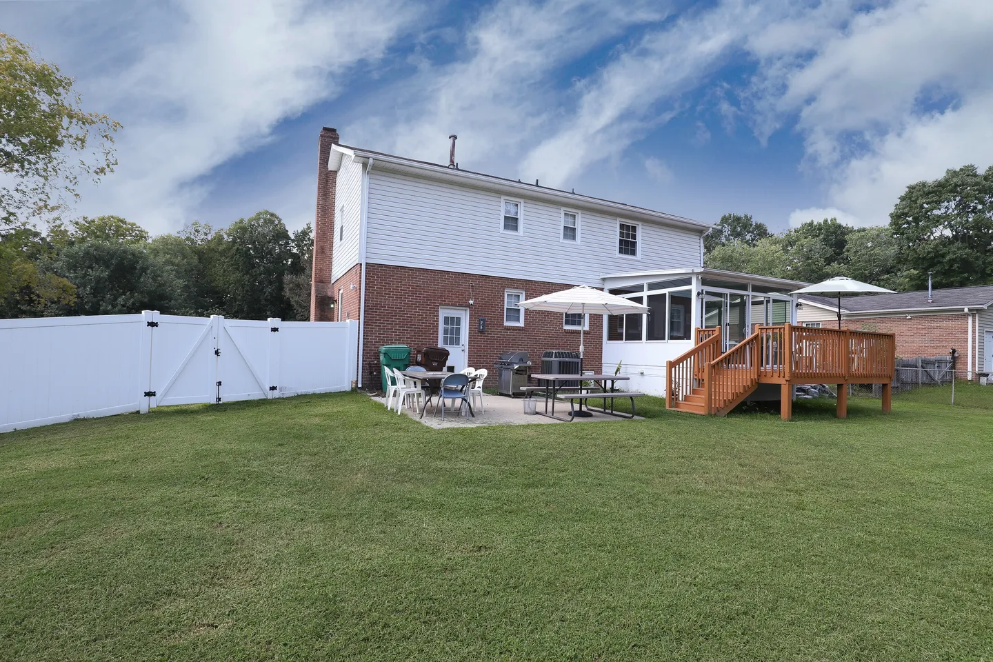Backyard of a two-story home with a green lawn, wooden deck, patio dining sets, screened porch, and white vinyl fence.