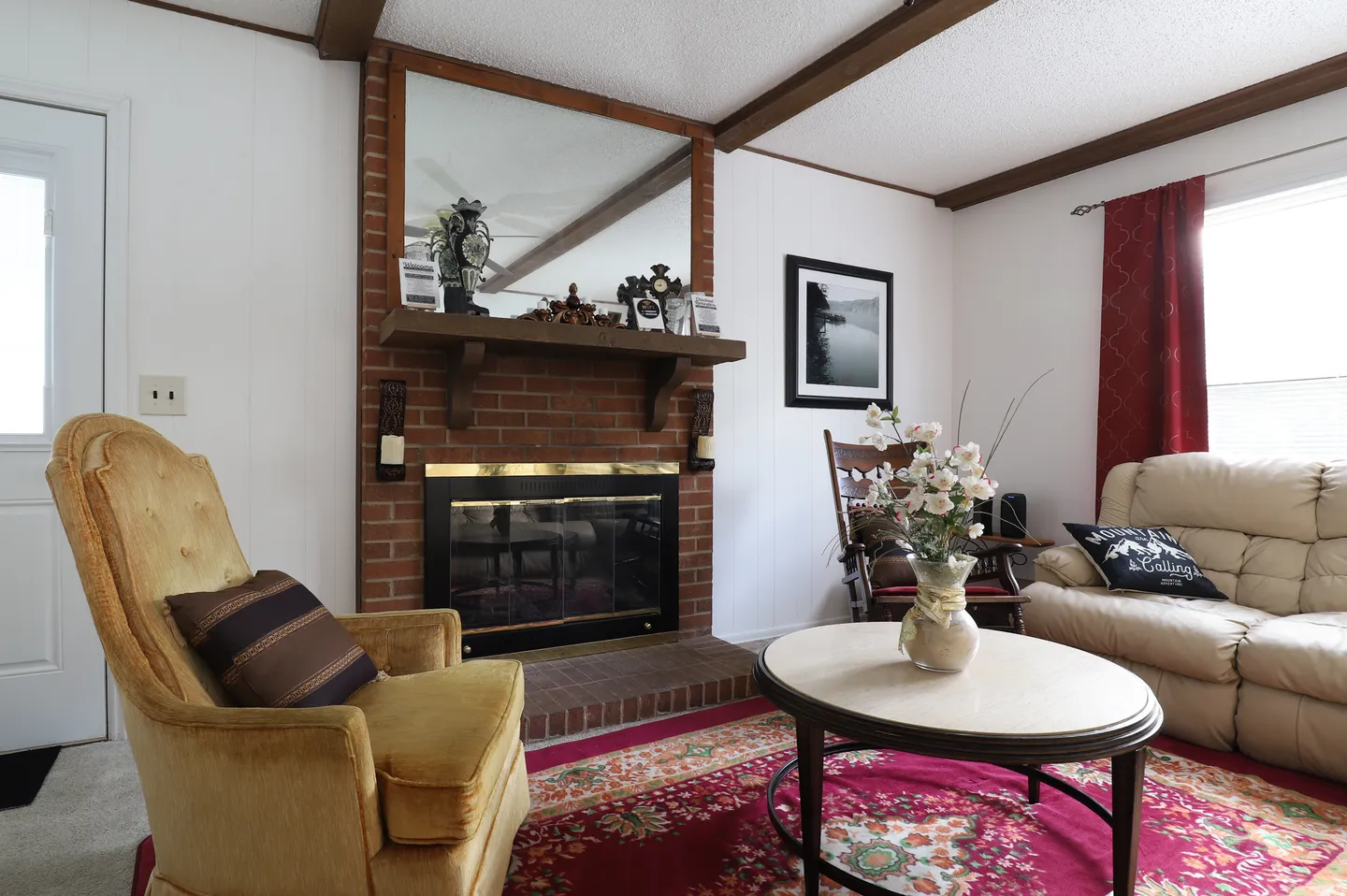 Living room with a red brick fireplace, wood mantel, large mirror, yellow armchair, round coffee table, and patterned rug.