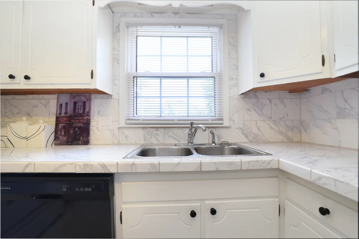 Kitchen counter with a double stainless steel sink beneath a window, surrounded by white tiled countertops and white cabinets.