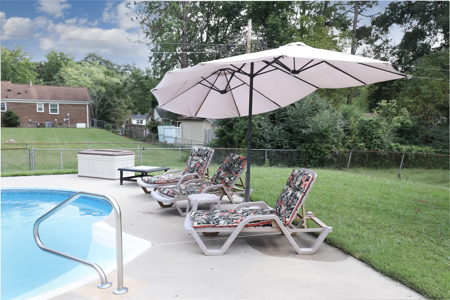 Outdoor poolside patio with lounge chairs under a large umbrella beside an in-ground swimming pool and grassy yard.