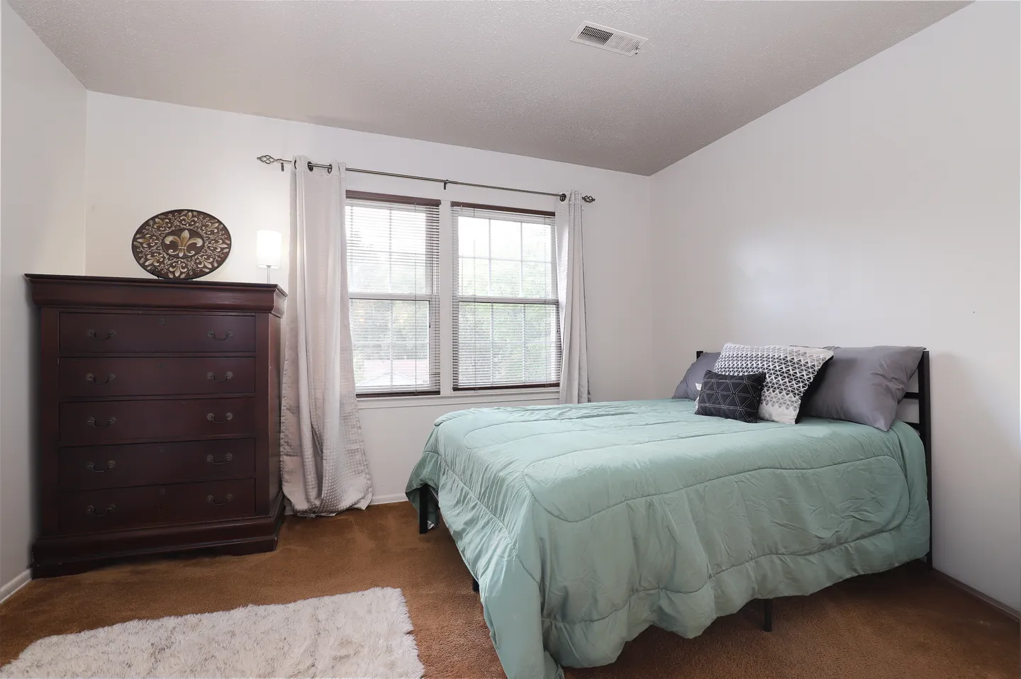 Bedroom featuring a made bed with a light green comforter, a dark wood dresser, carpeted flooring, and a double window with blinds and curtains.