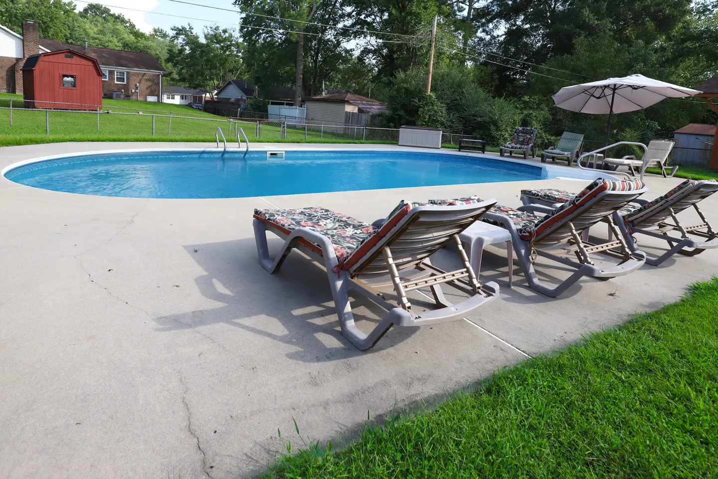 Outdoor swimming pool surrounded by a concrete patio, cushioned lounge chairs, and a grassy backyard with a red shed.