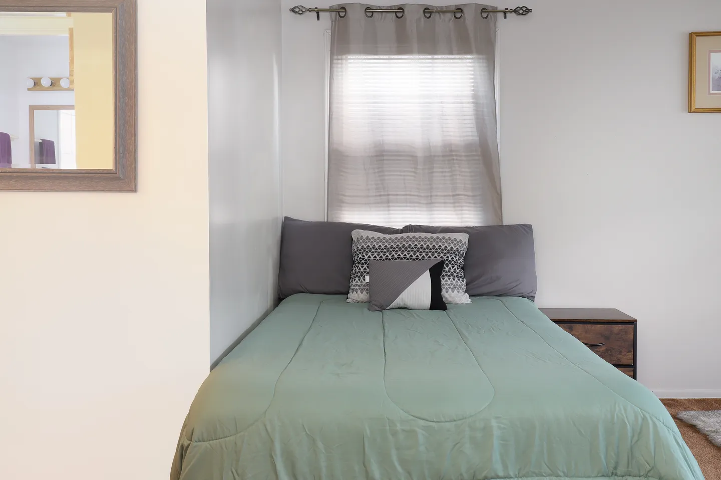 Bedroom nook with a bed made with a light green comforter, multiple accent pillows, and a window behind it.