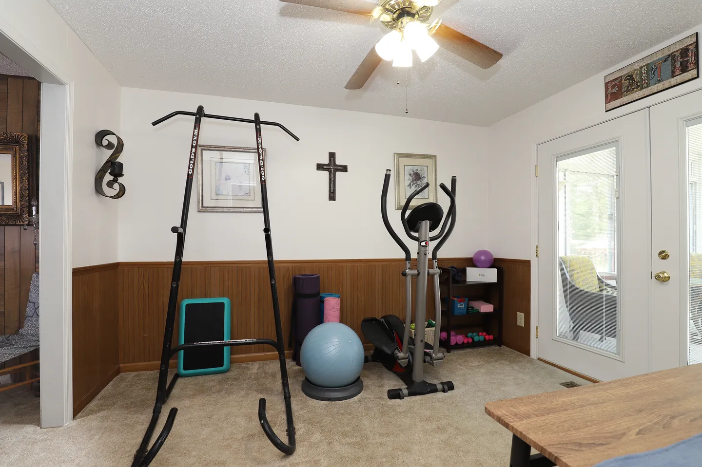 Home workout room with an elliptical, pull-up tower, exercise ball, and yoga accessories alongside carpeted flooring and wood wainscoting.
