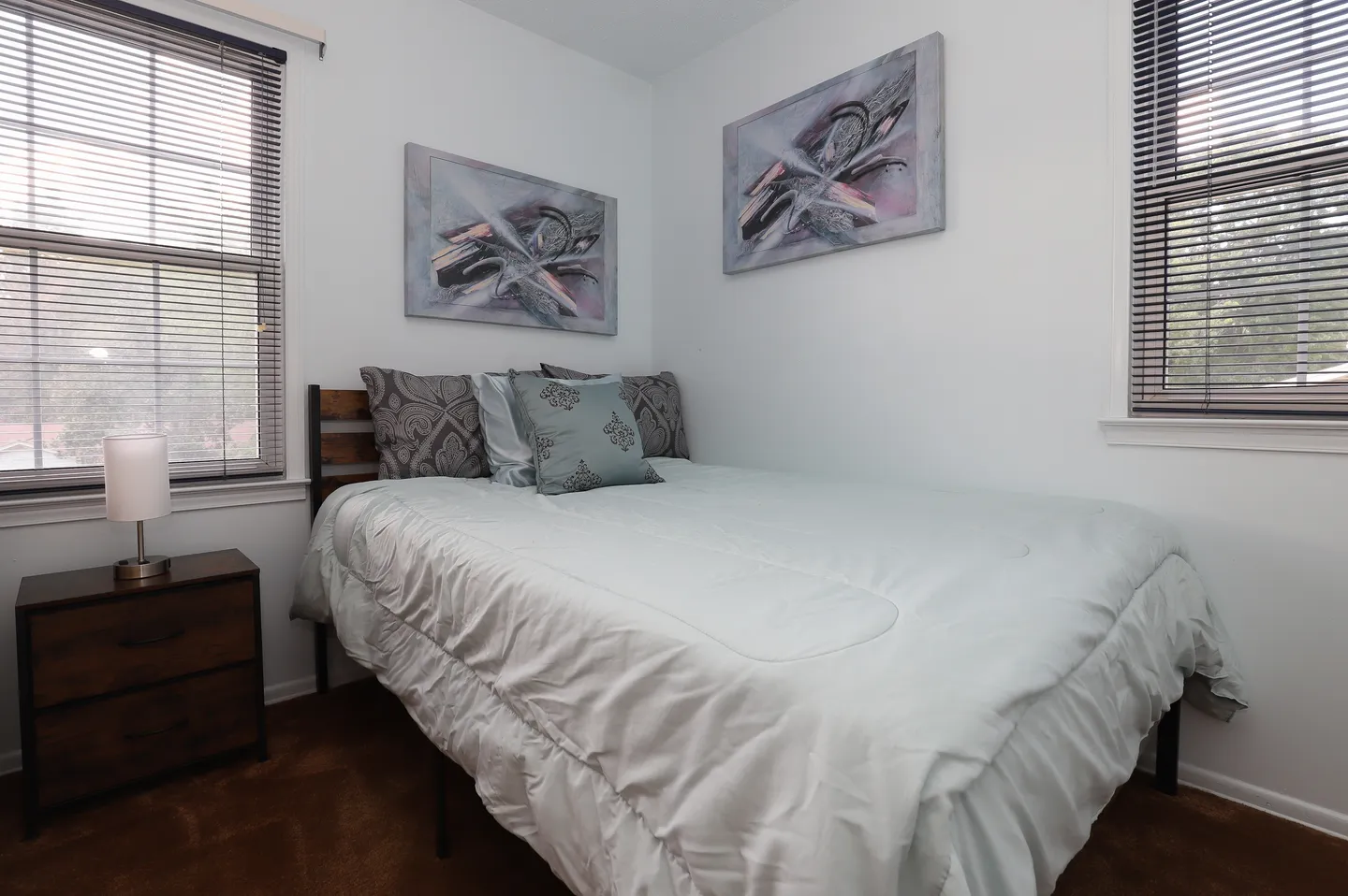 Bedroom with a bed made with light bedding, dark accent pillows, a bedside table with a lamp, and two windows with blinds.