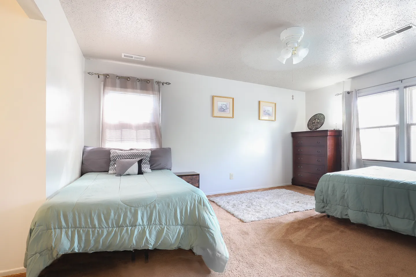 Bedroom featuring two beds made with light green comforters, tan carpeting, a ceiling fan, and a tall wooden dresser.