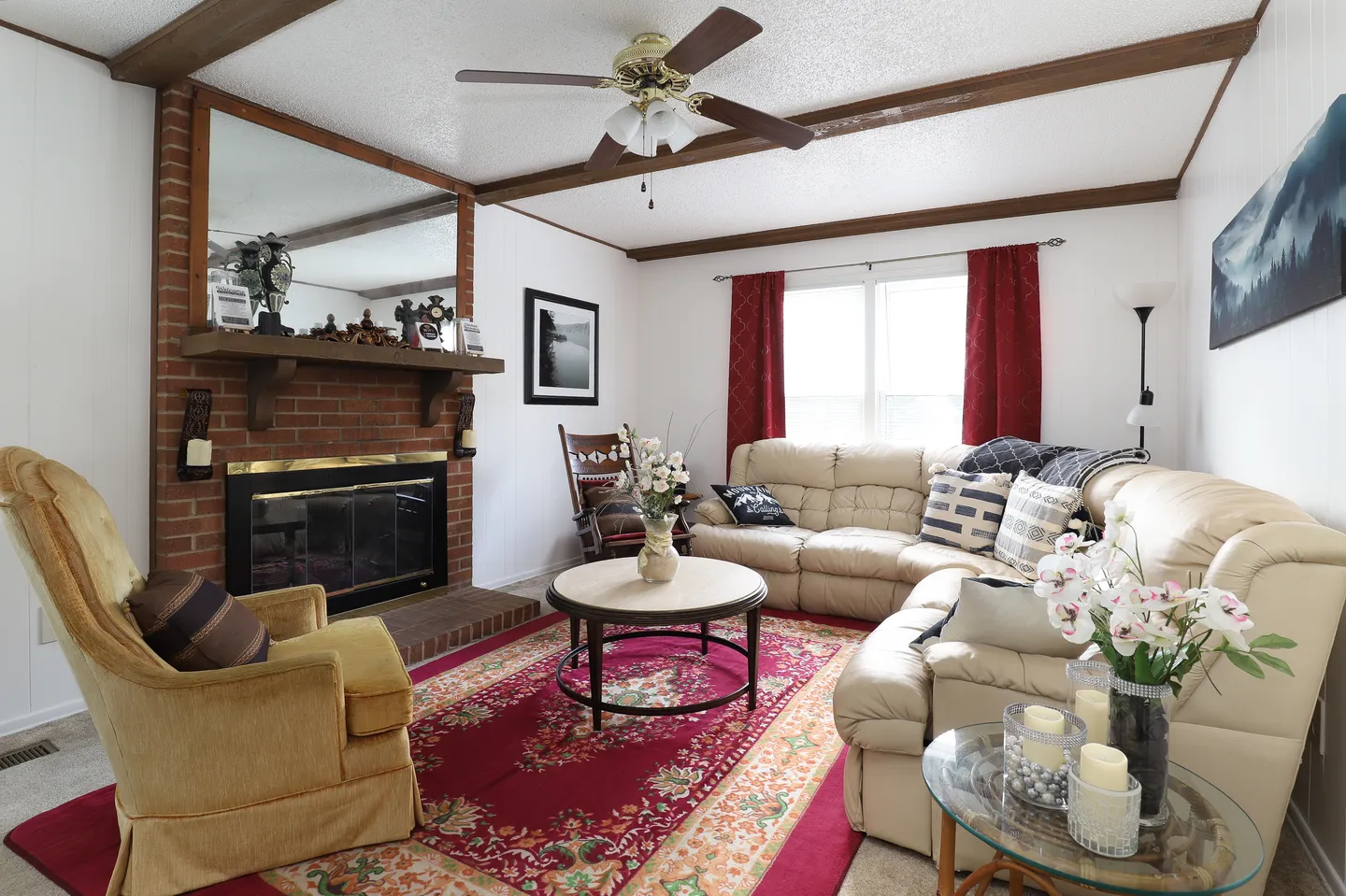 Living room with a brick fireplace, leather sectional sofa, ceiling fan, and a red patterned area rug.