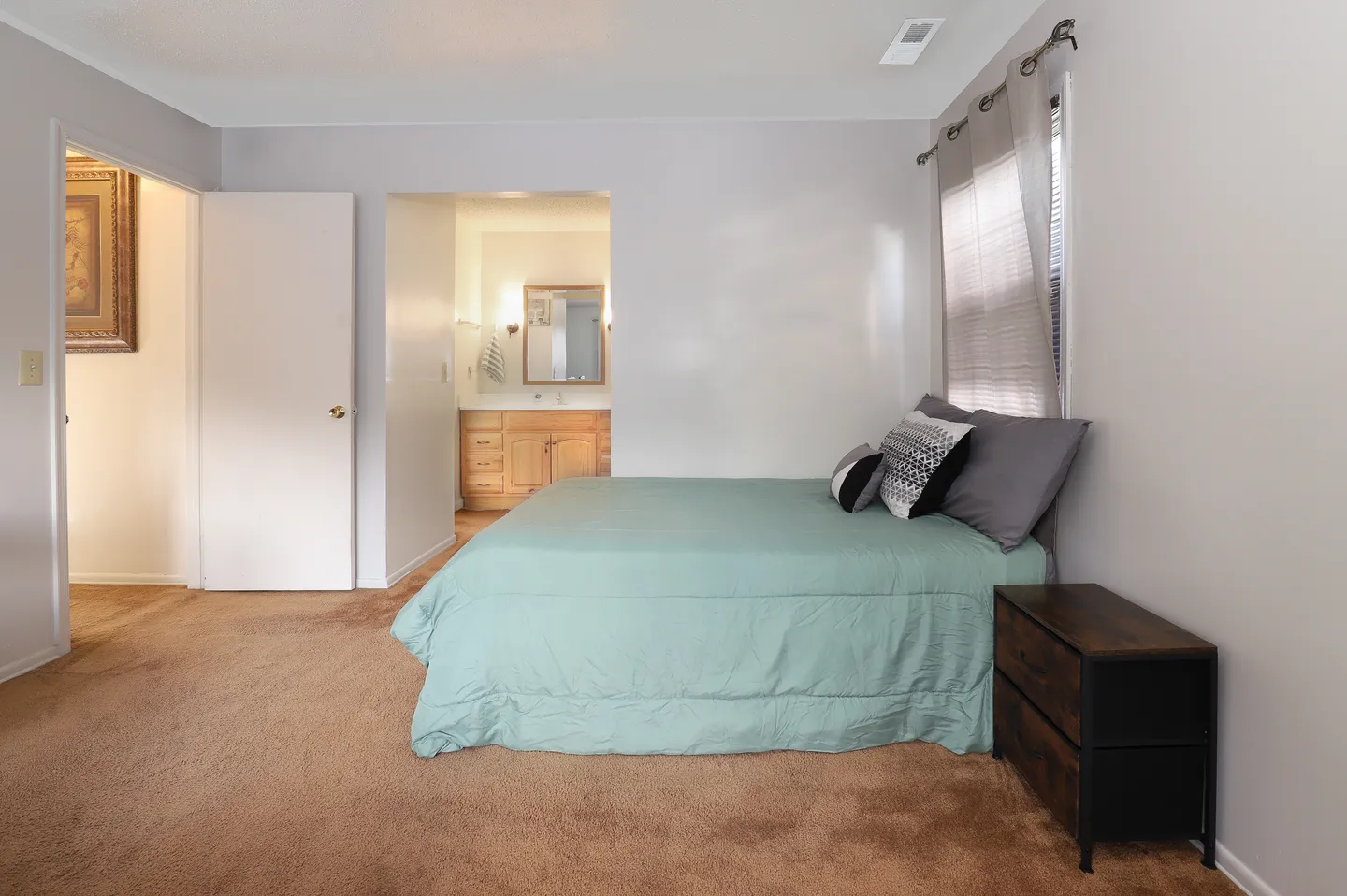 Bedroom with brown carpet, light teal bedspread, nightstand, and an ensuite bathroom entrance.