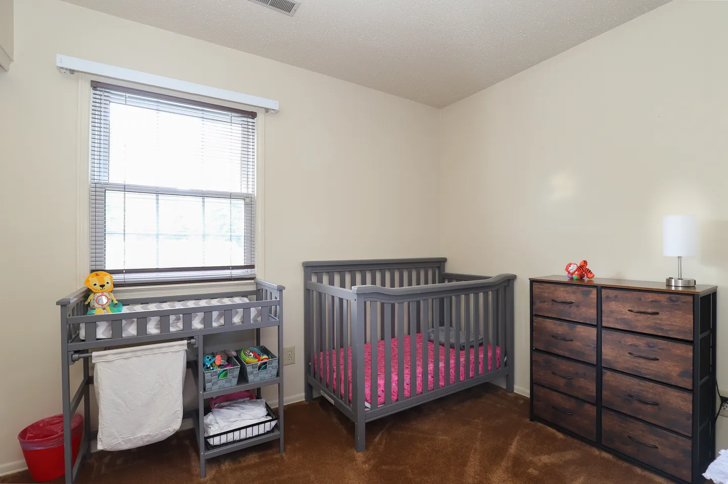 Nursery bedroom featuring a gray wooden crib, changing table, and a dark wood dresser against beige walls with brown carpet.