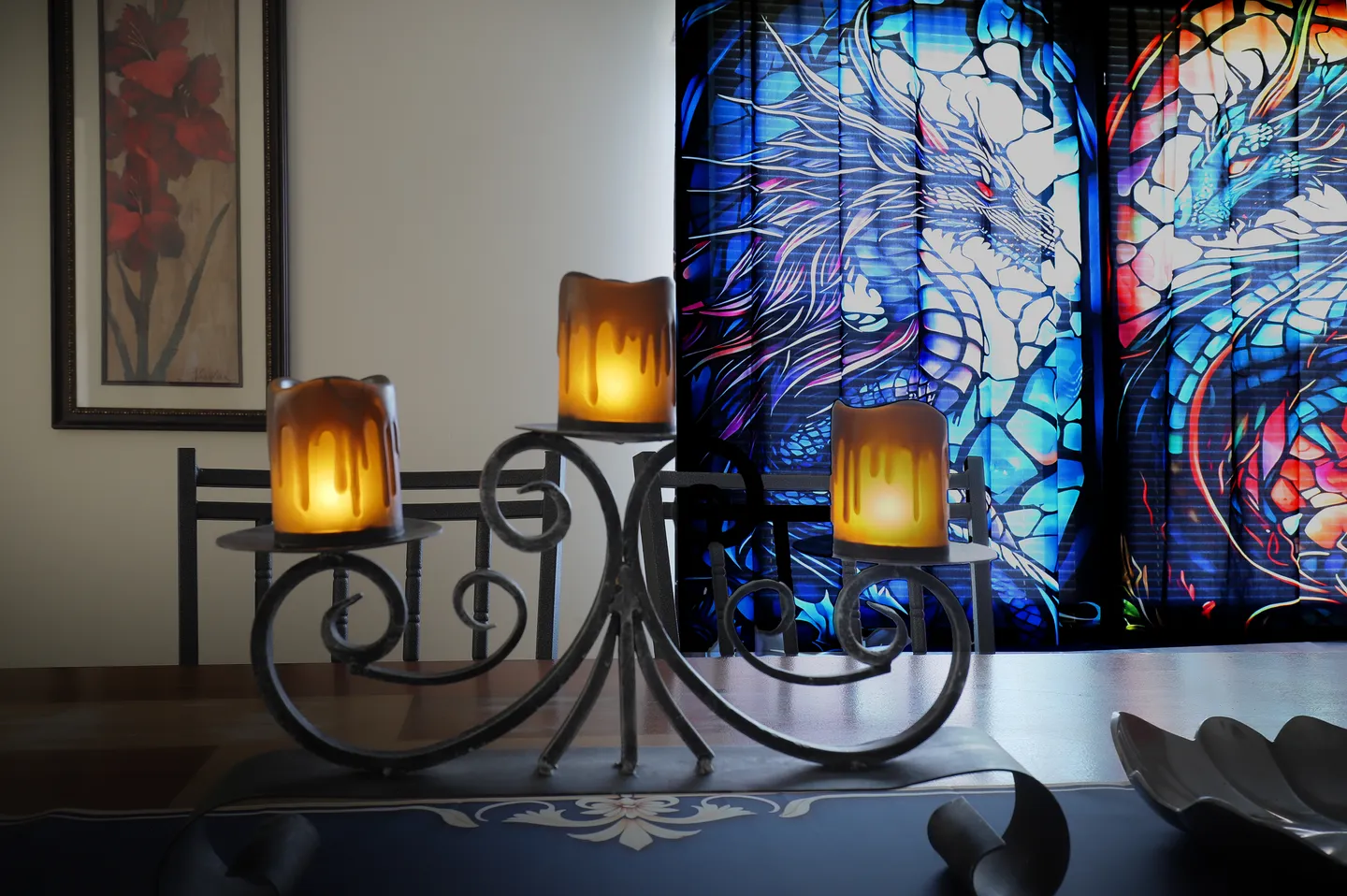 A wrought iron candelabra with three illuminated amber candle holders on a tabletop in front of vibrant stained-glass style dragon window blinds.