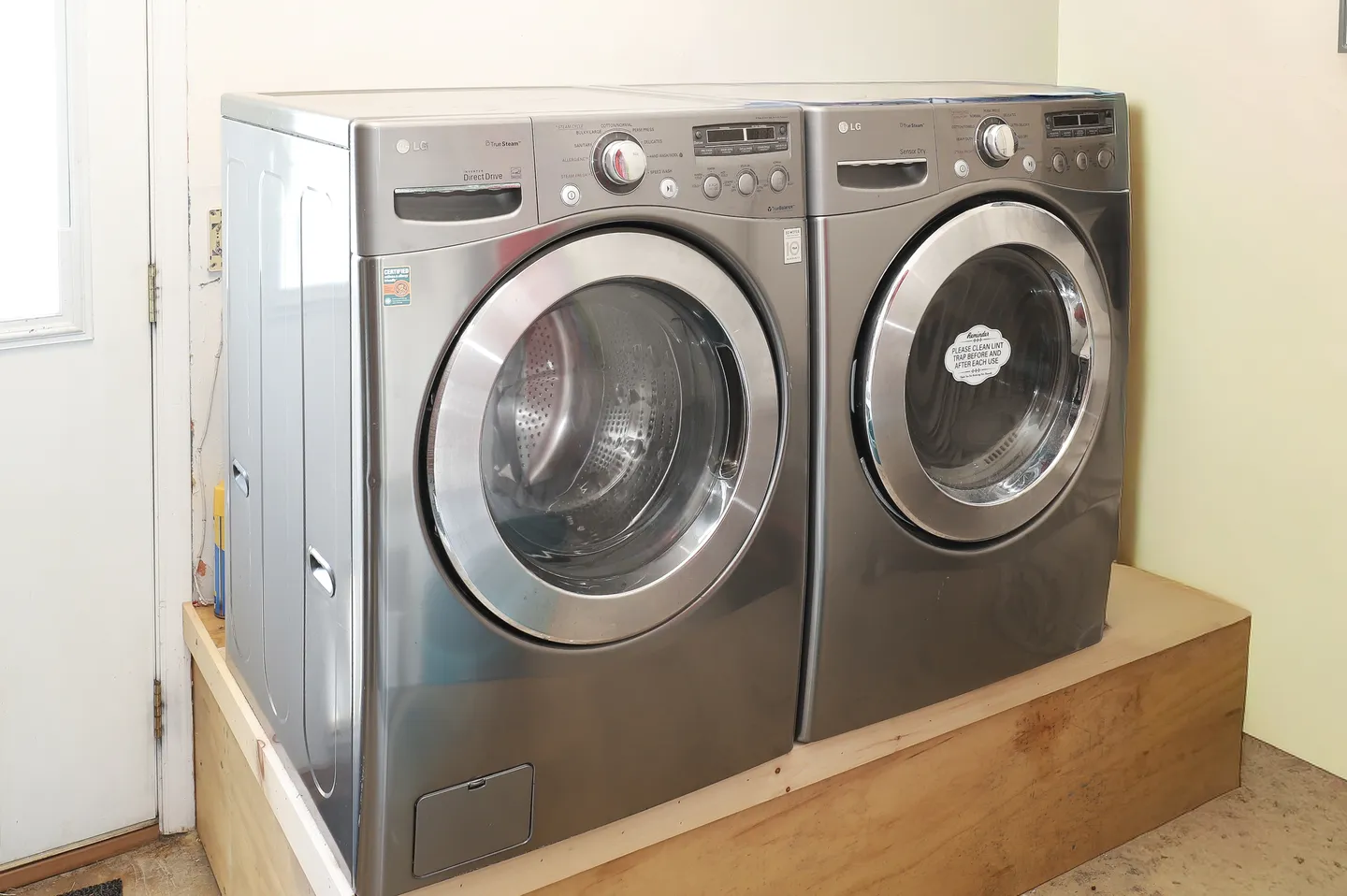 Front-loading metallic washing machine and dryer set side by side on a raised wooden platform indoors.