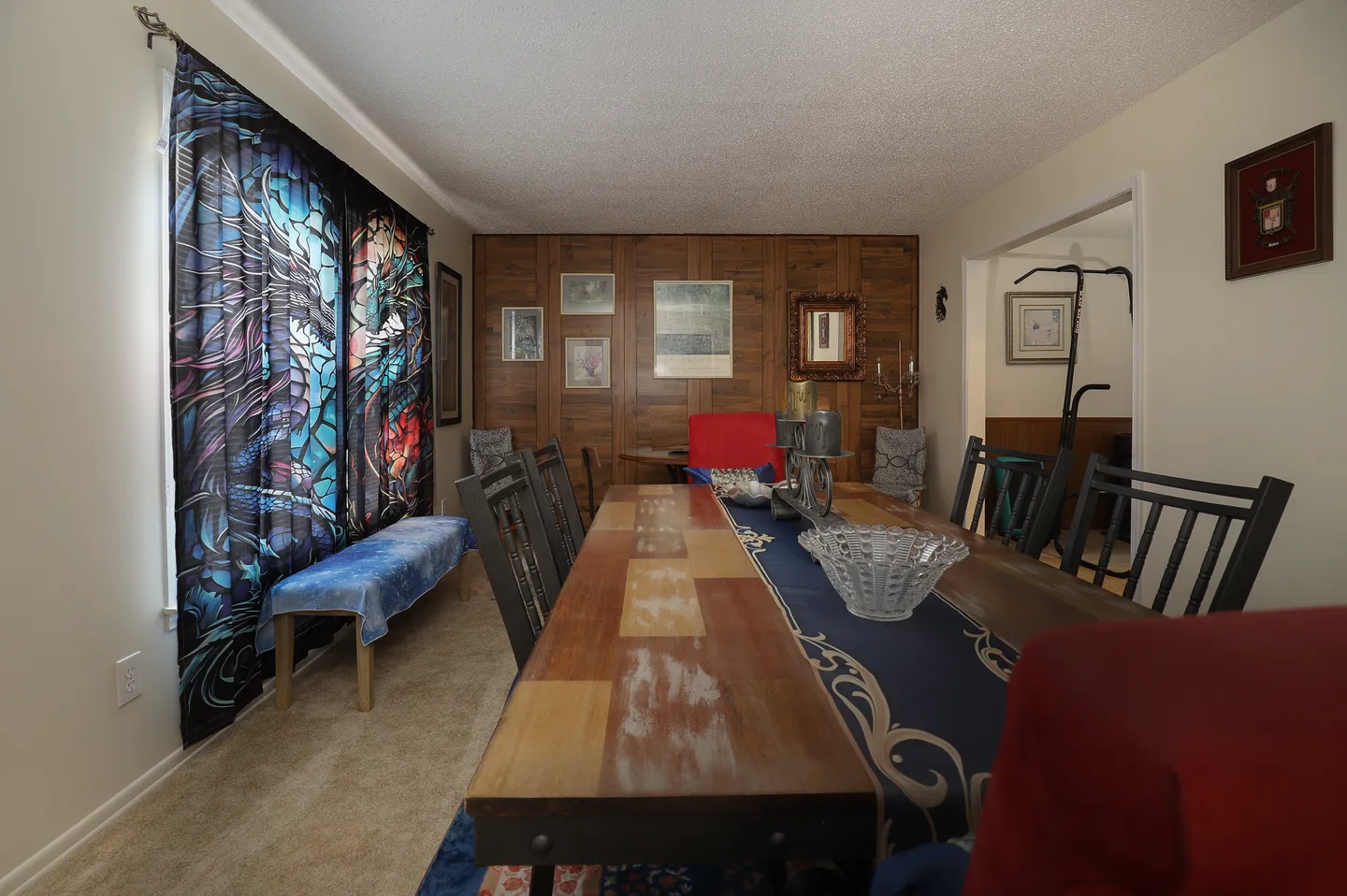 Dining area featuring a long wooden table with dark chairs, carpeted floor, paneled accent wall, and dragon-patterned curtains.