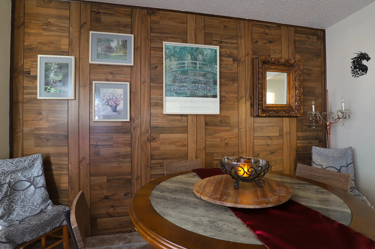 Dining area featuring a round table with chairs set against a wood-paneled wall decorated with framed art and a mirror.