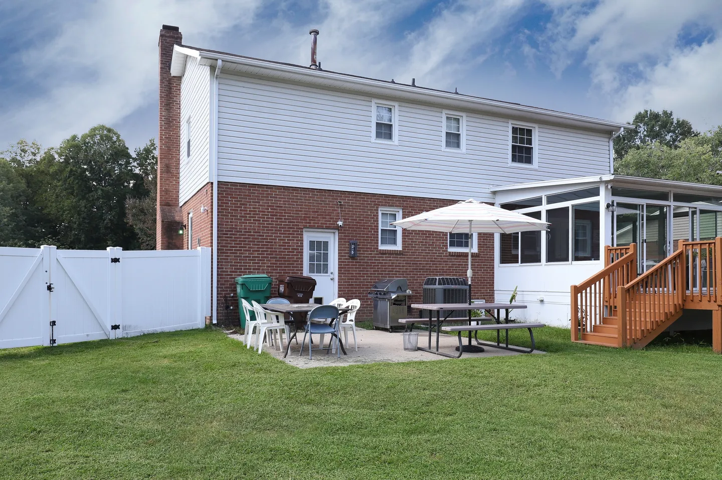 Rear exterior of a two-story home with a brick lower level, white siding upper level, patio with outdoor dining furniture, and wooden deck stairs.