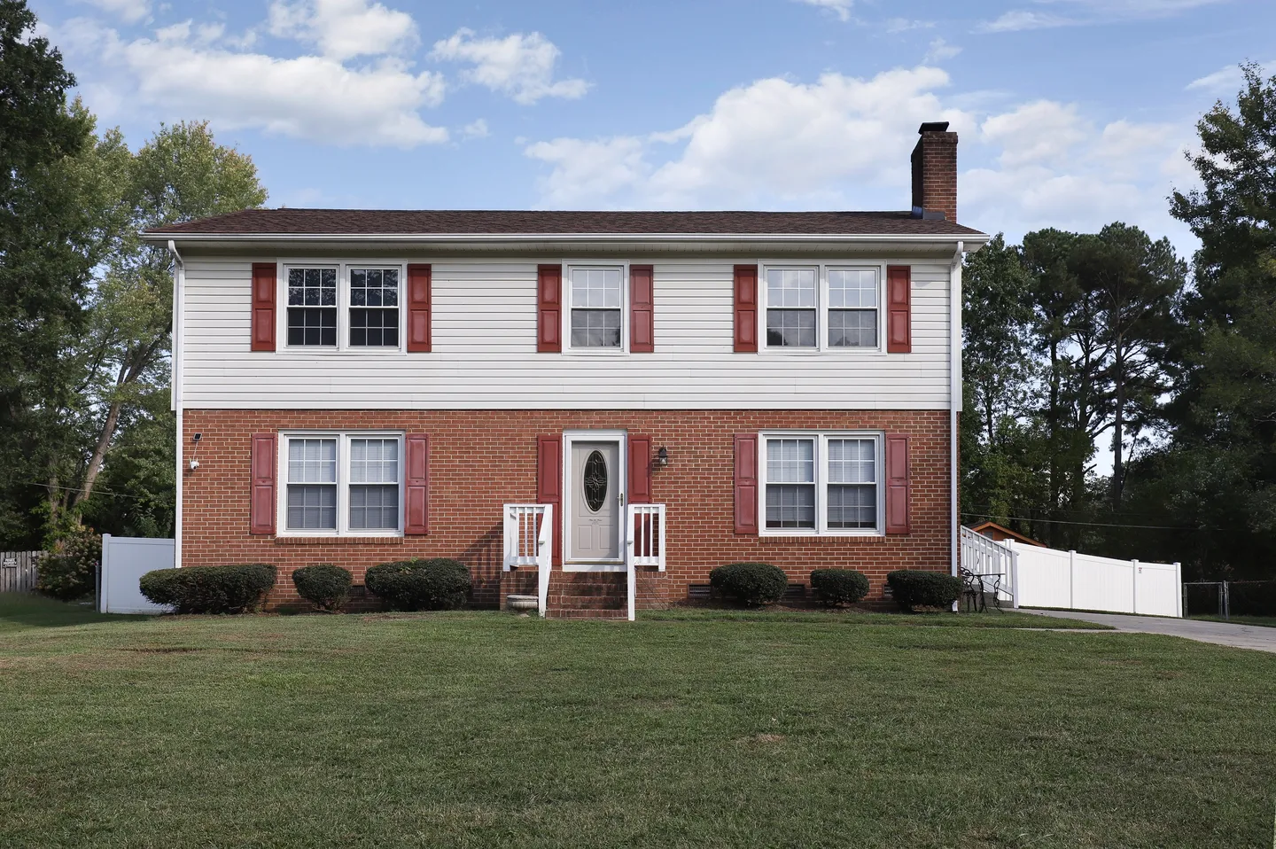 Front exterior of a two-story home with red brick on the lower level, white siding above, and red shutters, situated behind a front lawn.