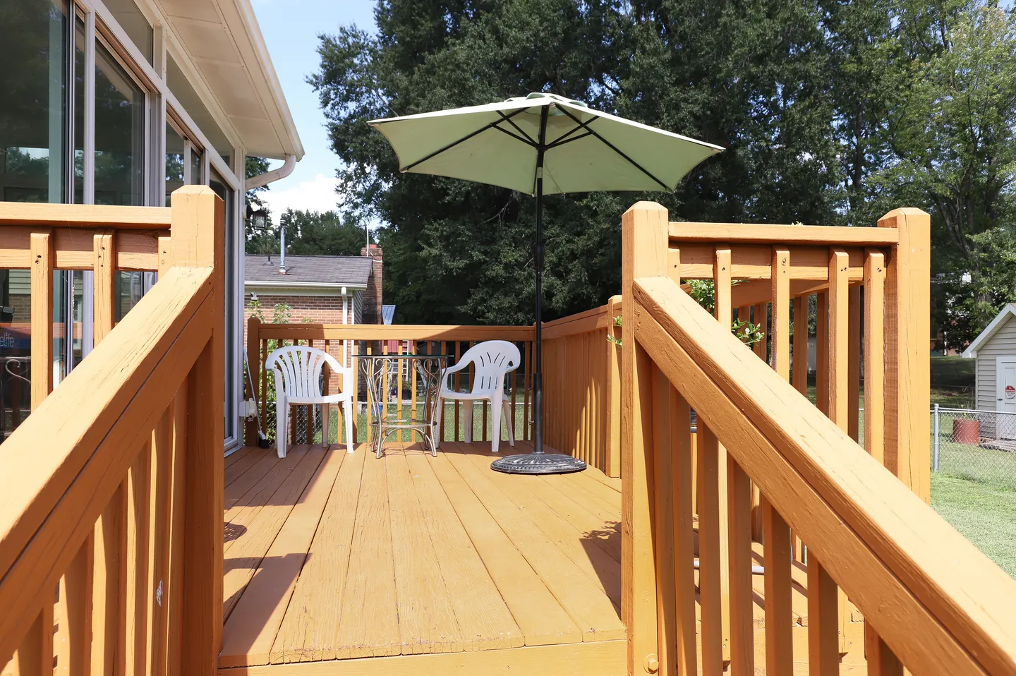 A wooden outdoor deck with stairs, a patio table, white chairs, and an umbrella outside sliding glass doors.