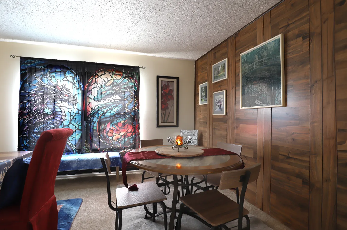 Dining area featuring a round table with chairs against a wood-paneled accent wall and large window with printed dragon curtains.