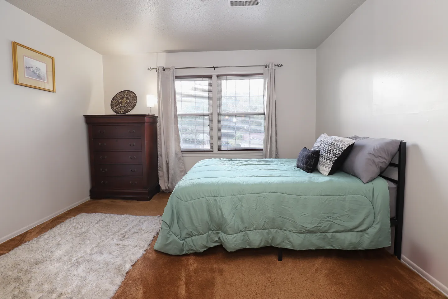 Bedroom featuring a made bed with a light green comforter, a tall dark wood dresser, a window with curtains, and carpeted floors.