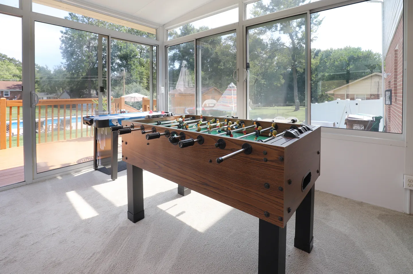 Sunroom with carpeted floor, foosball table, air hockey table, and glass windows overlooking an outdoor deck and pool.