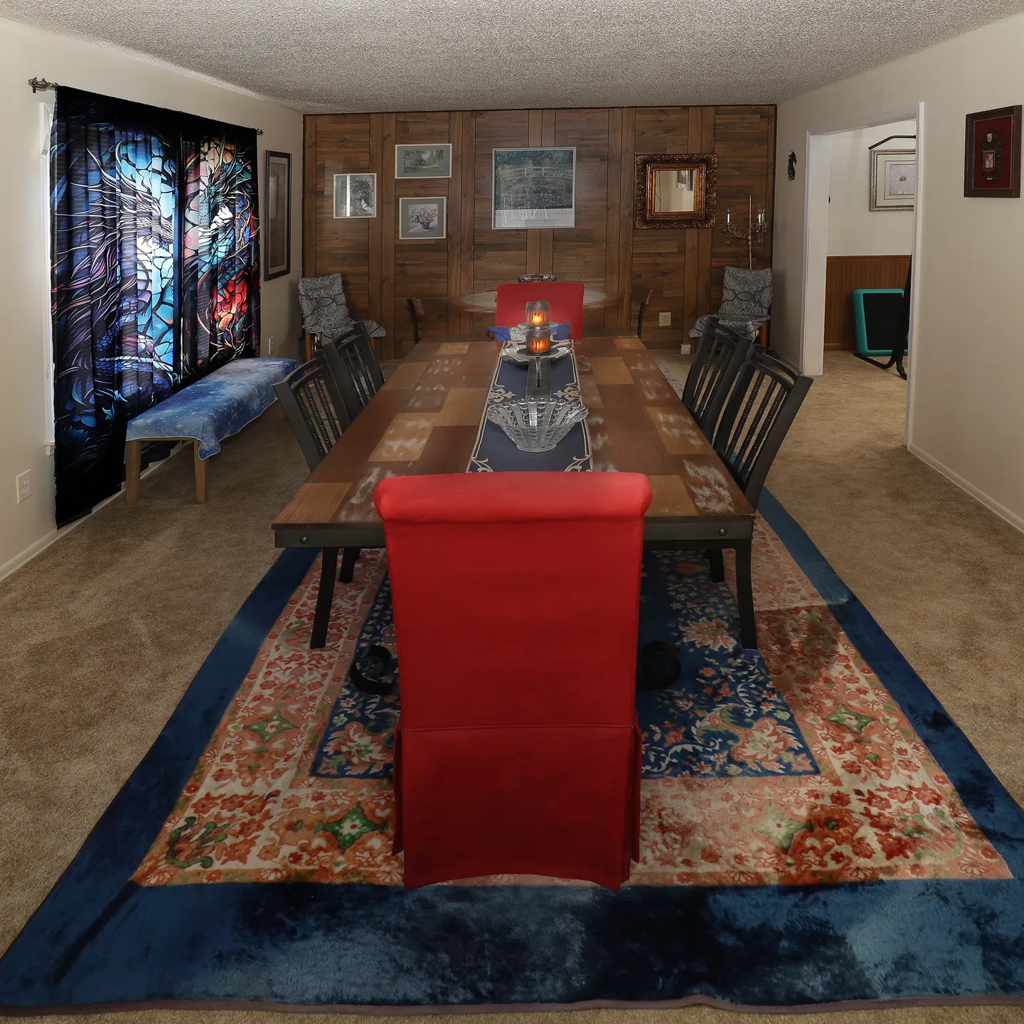 Dining room with a long table, carpeted flooring, wood-paneled accent wall, and decorative window curtains.