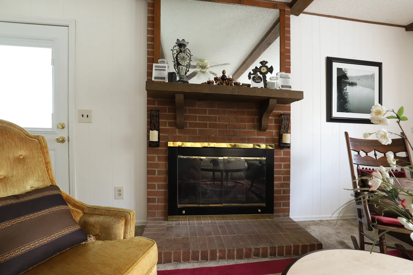 Red brick fireplace with a wood mantel and large mirror above, flanked by white paneled walls and seating in a living room.