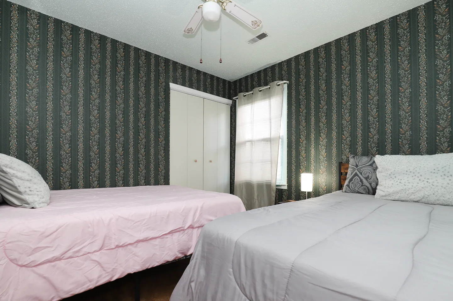 Bedroom featuring two beds, dark patterned wallpaper, a curtained window, and a ceiling fan.