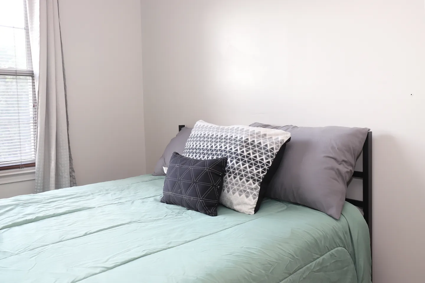 A made bed with a light green comforter, patterned accent pillows, and a black metal frame next to a window with blinds.
