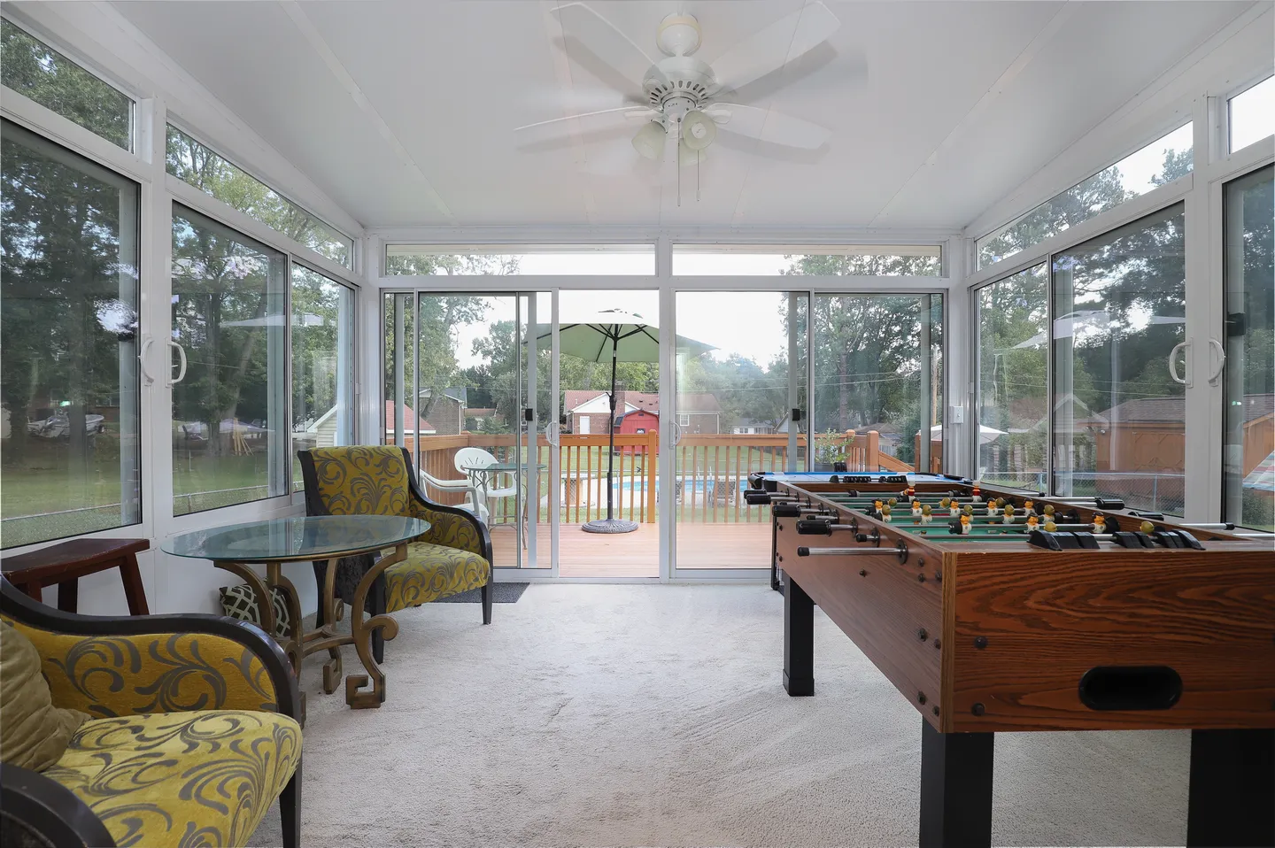 Sunroom with wall-to-wall windows, a foosball table, armchairs around a small glass table, and sliding doors leading to an outdoor deck.
