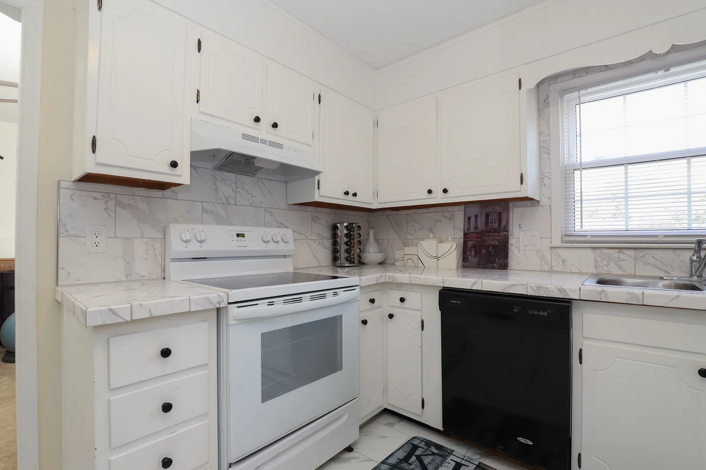 Kitchen corner showing white cabinets, white electric stove, black dishwasher, and marble-look tiled countertops.