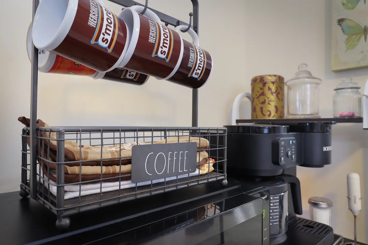 Close-up of a coffee station featuring a wire basket labeled 'COFFEE', hanging mugs, and a black Keurig coffee maker.