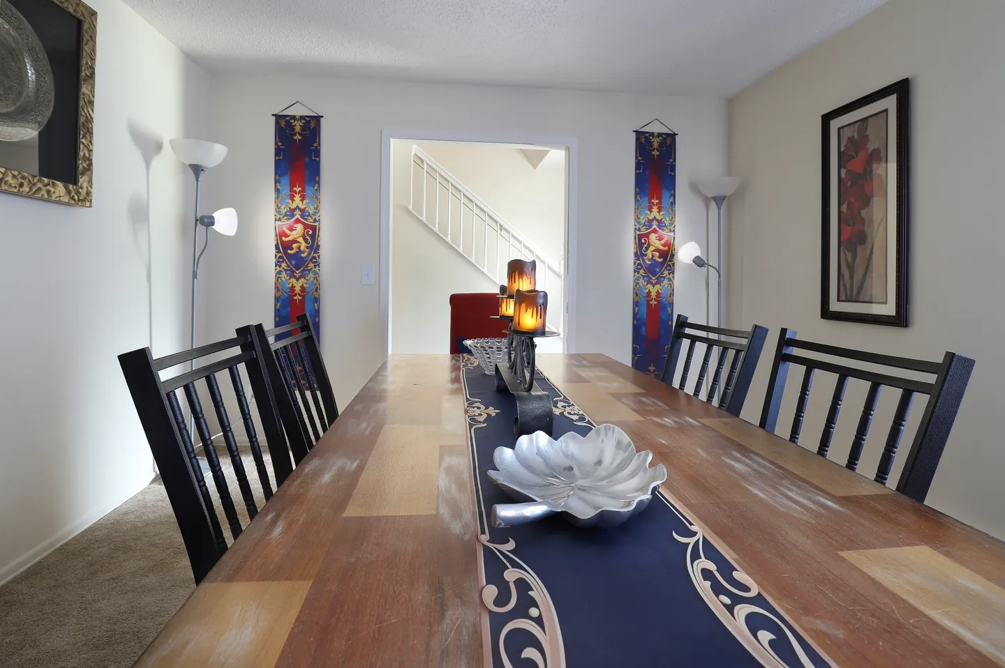 Dining area with a long wooden table flanked by dark slatted chairs, decorative house banners on the wall, and a doorway leading toward a staircase.