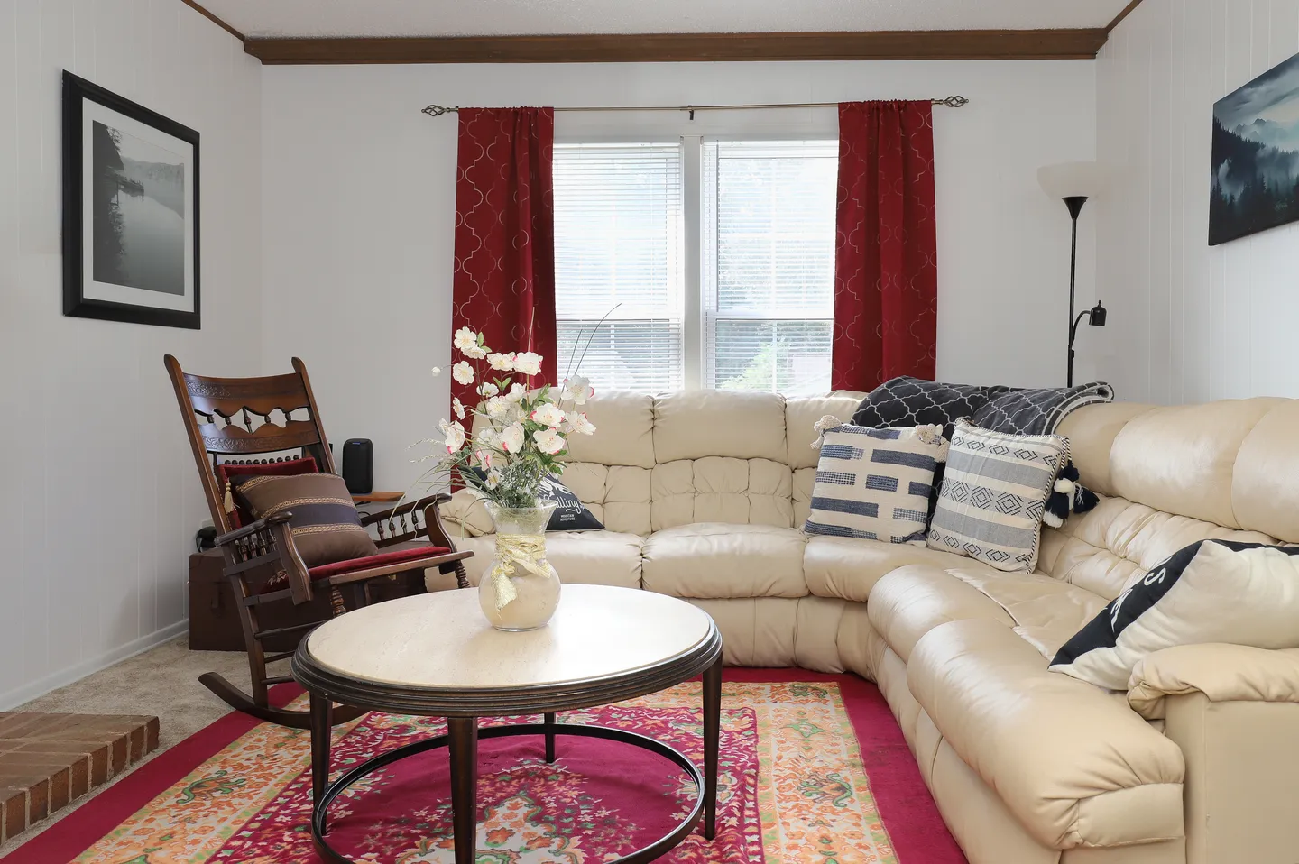 Living room with a large cream-colored sectional sofa, a round coffee table, a wooden rocking chair, and a bright red patterned rug.