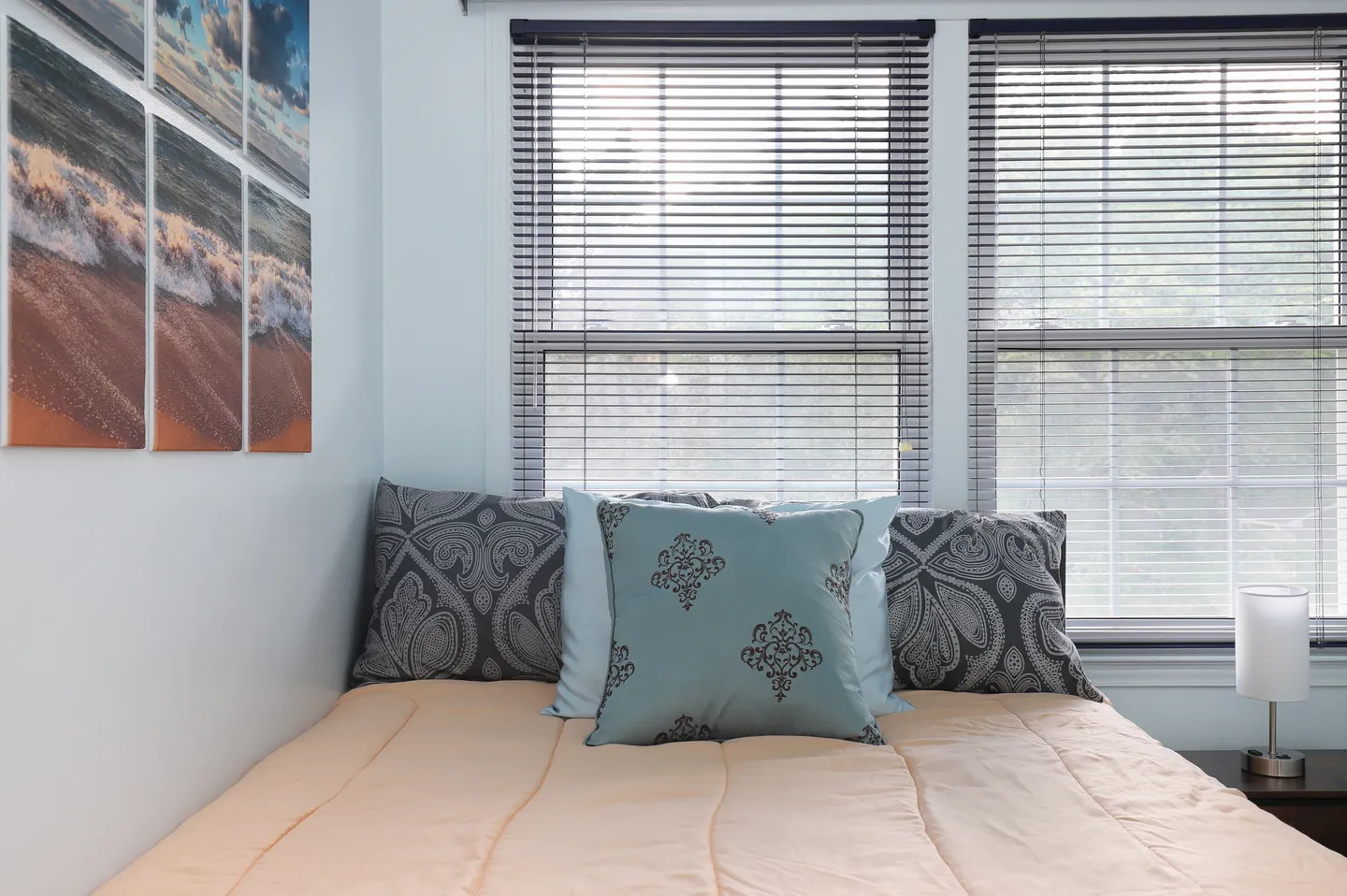 A made bed tucked into a corner beneath two windows with blinds, decorated with patterned throw pillows and ocean wall art.