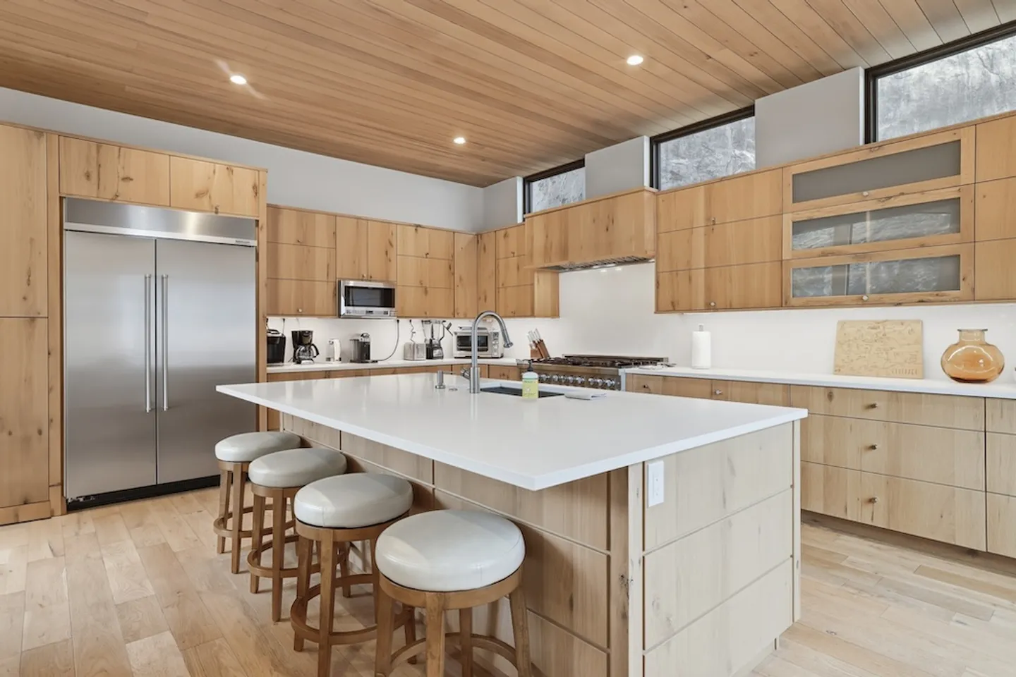 Modern kitchen with light wood cabinets, wood-paneled ceiling, large island with barstools, and stainless steel appliances.