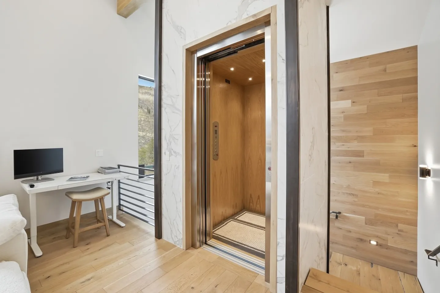 Open residential elevator with wood-paneled interior beside a small workspace with a desk and monitor.