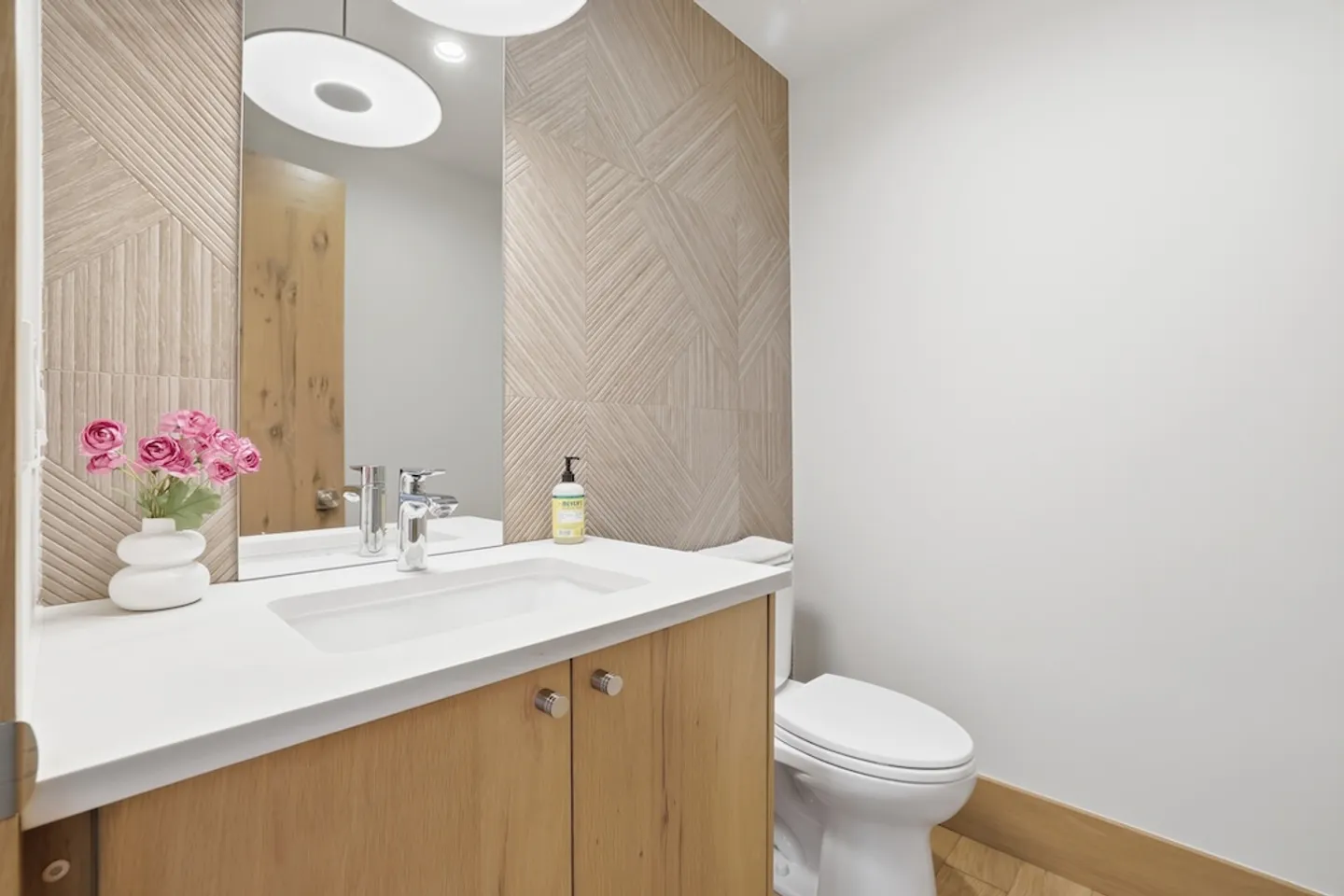 Bathroom with light wood vanity, white countertop, geometric accent wall tile, mirror, and white toilet.