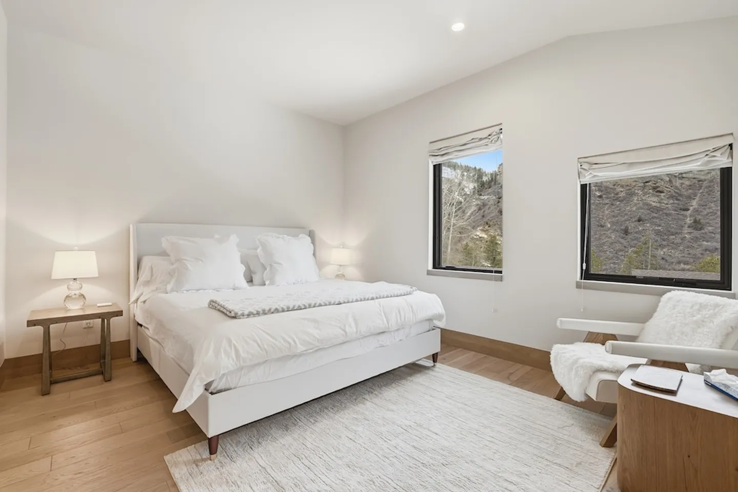 Bright bedroom with a white upholstered bed, light wood floors, and two windows offering views of a mountainside.