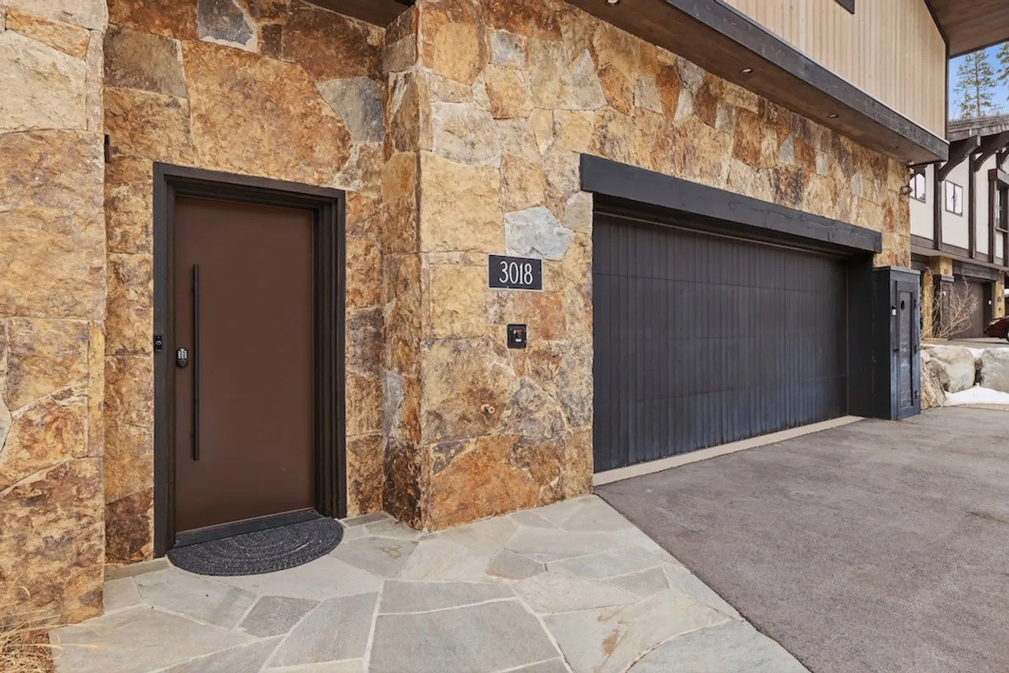 Exterior ground-level entrance with stone facade, brown entry door with vertical handle, and adjacent dark garage door.