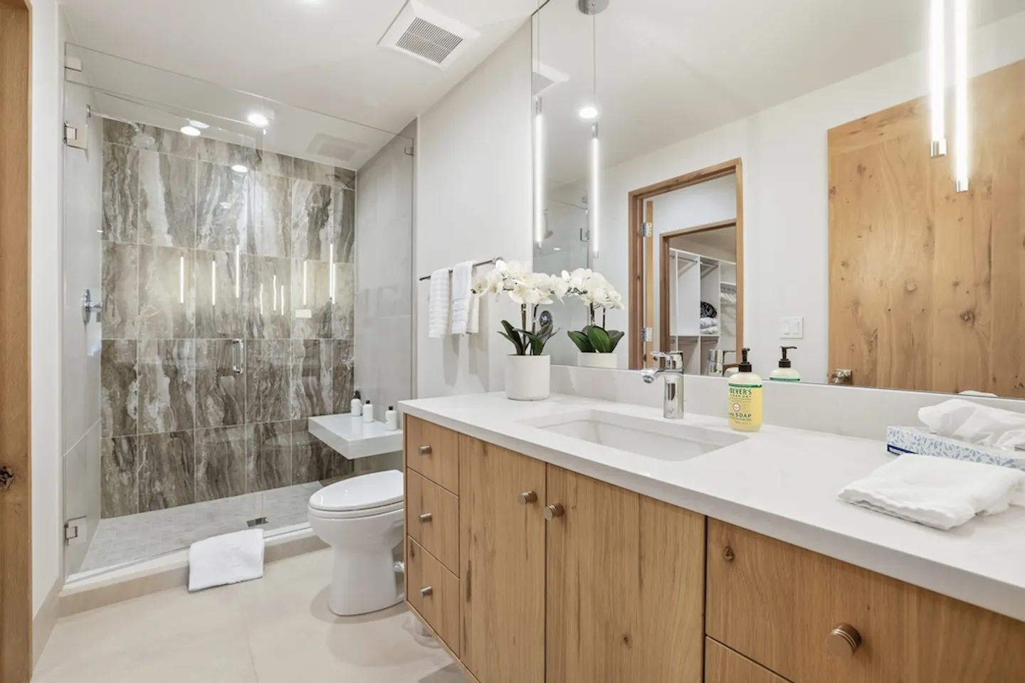 Bathroom vanity with light wood cabinetry, white countertop, and a walk-in glass shower tiled in patterned stone.