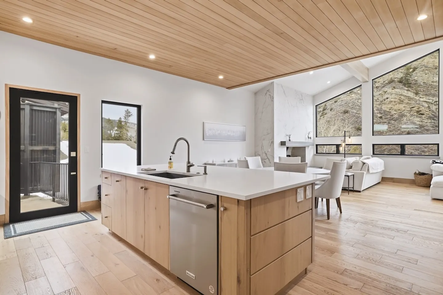 Open-concept kitchen island with a light wood base, integrated sink, and dishwasher, leading into a bright dining and living area.