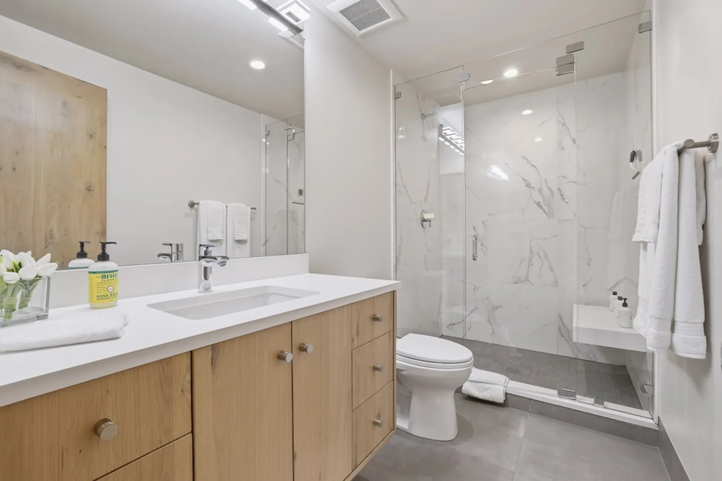 Bathroom with light wood vanity, white countertop, large mirror, toilet, and a glass-enclosed walk-in shower with marble-look tiles.