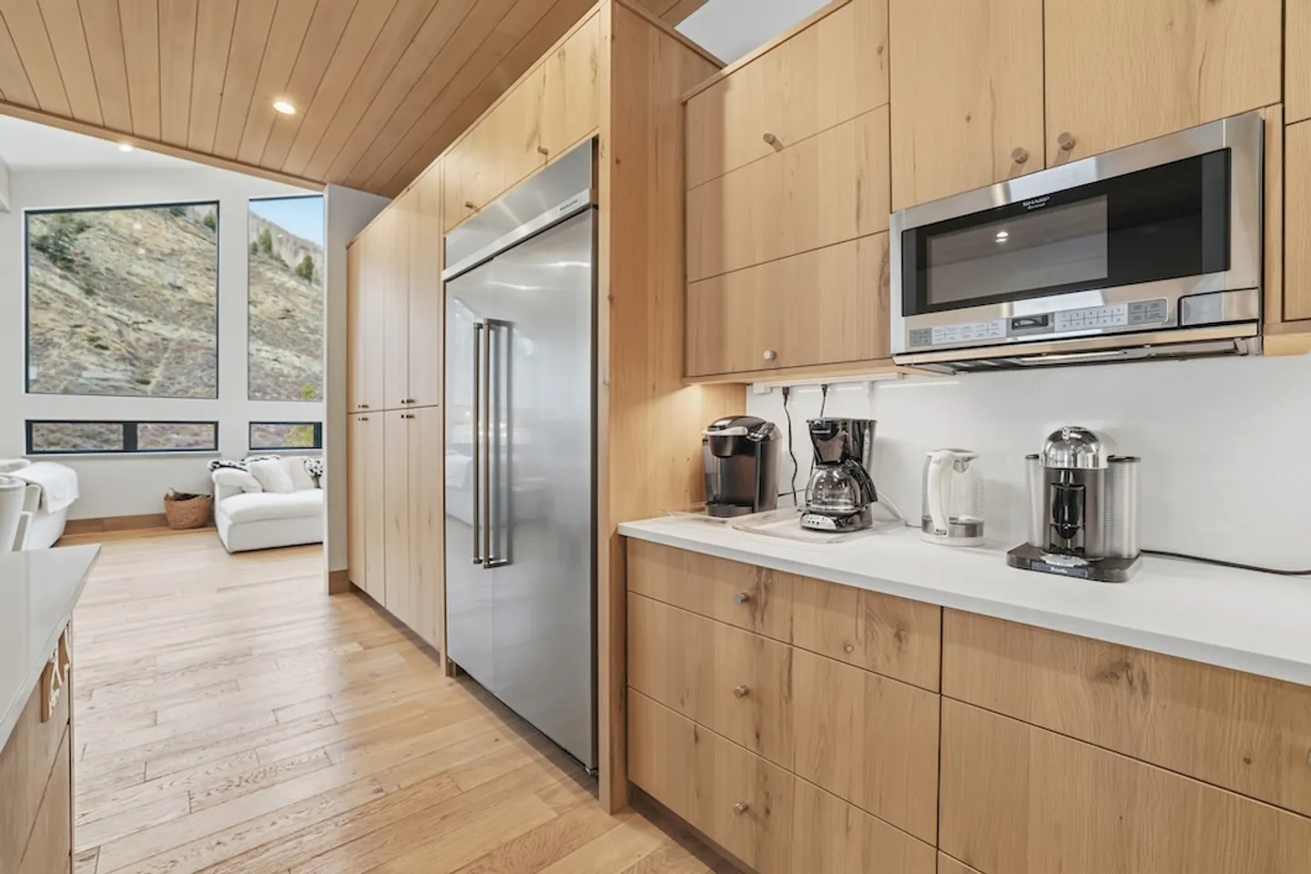 A modern kitchen with light wood cabinetry, stainless steel refrigerator, microwave, and small appliances on the white countertop.
