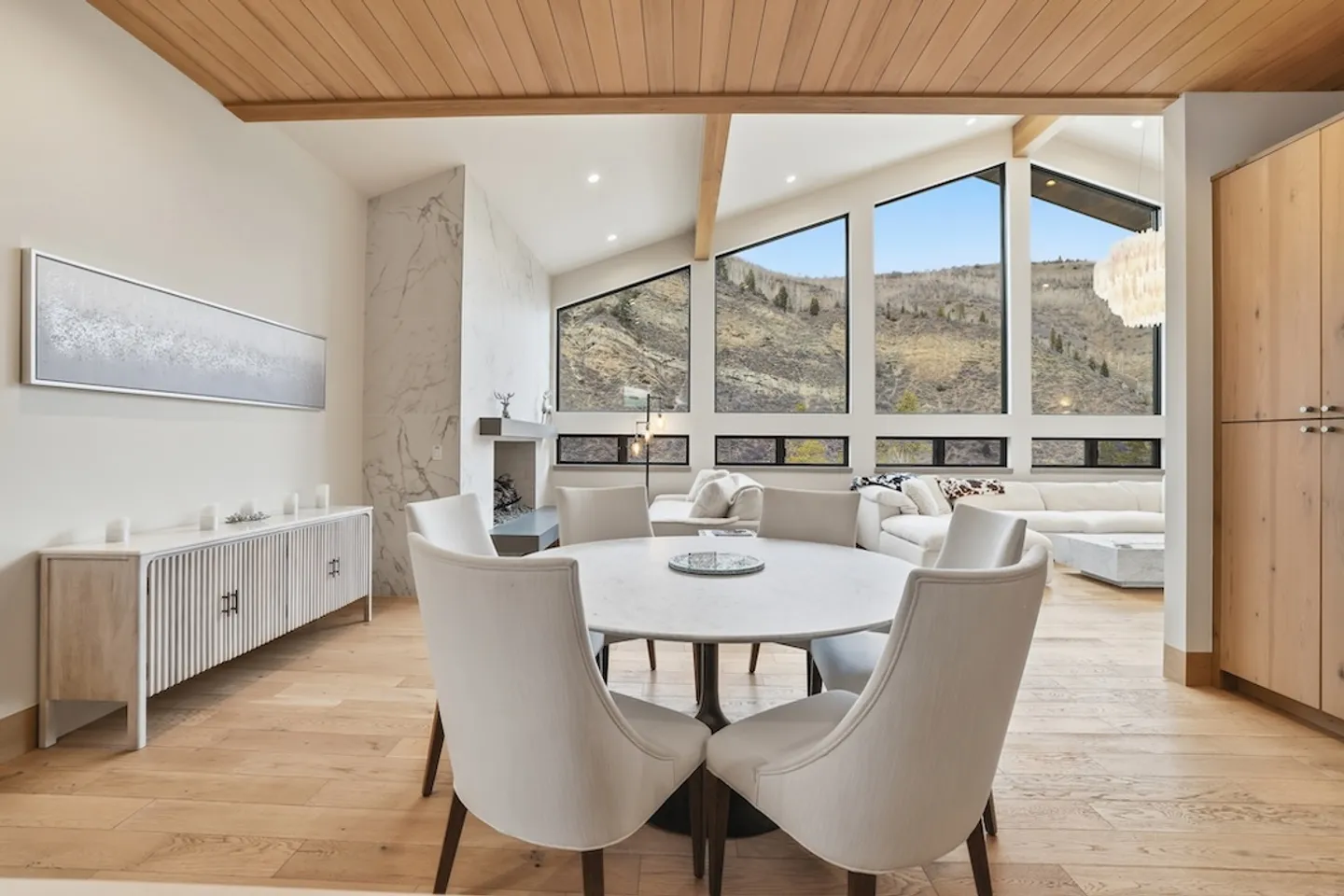 Open-concept dining and living area with a round dining table, wood-plank ceiling, and large windows framing mountain views.