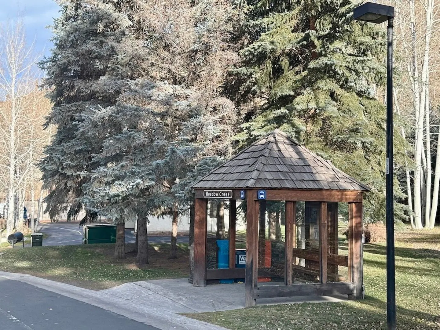 A wooden gazebo-style bus stop shelter labeled Meadow Creek beside a paved road and pine trees.