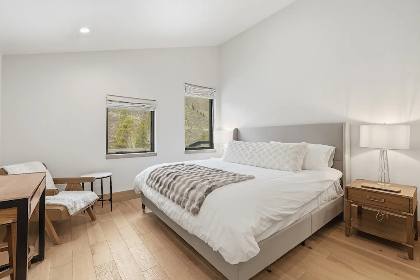 Bedroom with light wood flooring, an upholstered bed with white bedding, nightstand with lamp, and an armchair near two windows overlooking hills.