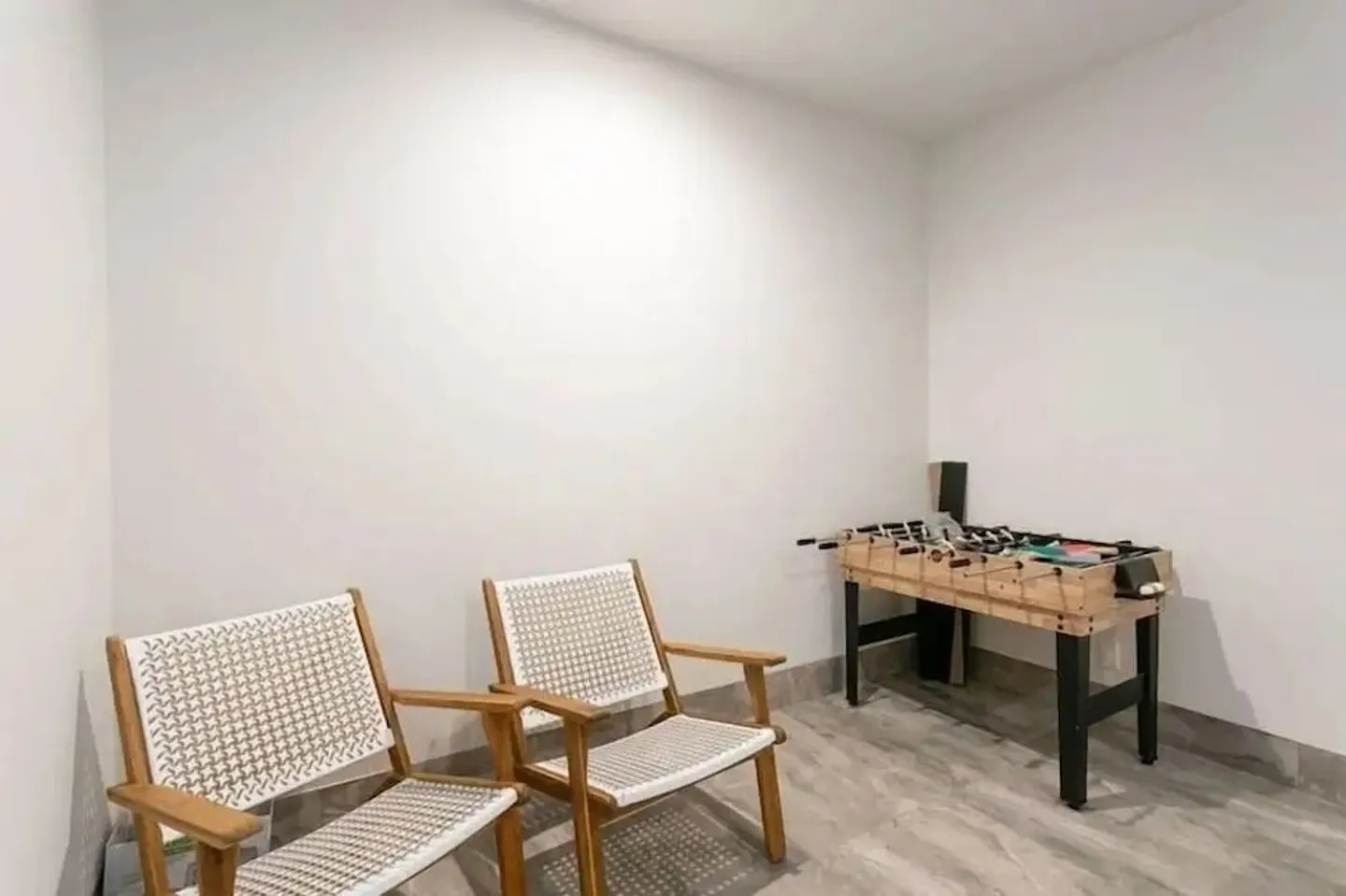 Indoor game area with white walls, tiled flooring, two wooden chairs with woven seats, and a foosball table.
