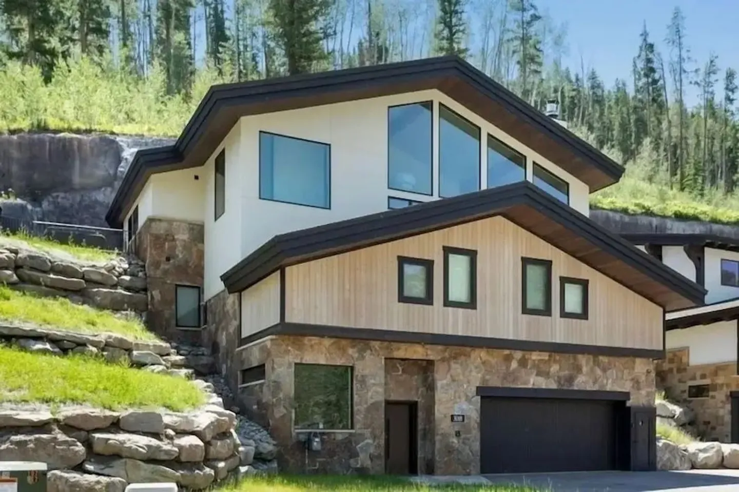 Exterior front view of a multi-level modern mountain home with stone siding, light wood accents, and a tiered rock landscaping wall.