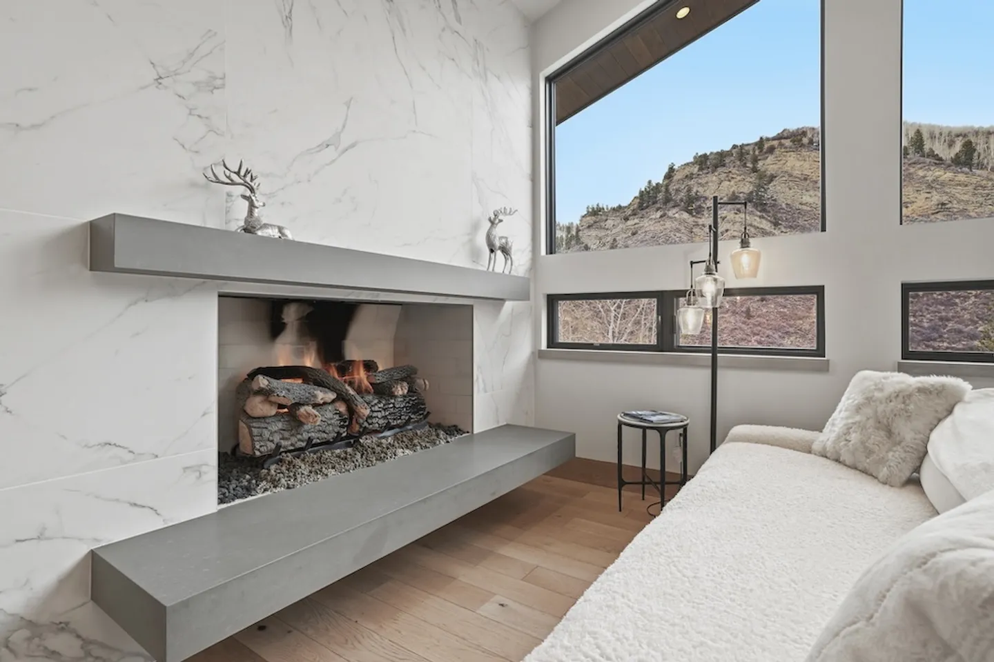 A modern marble-look fireplace with a lit fire, gray mantle, textured white sofa, and large windows revealing a mountain view.
