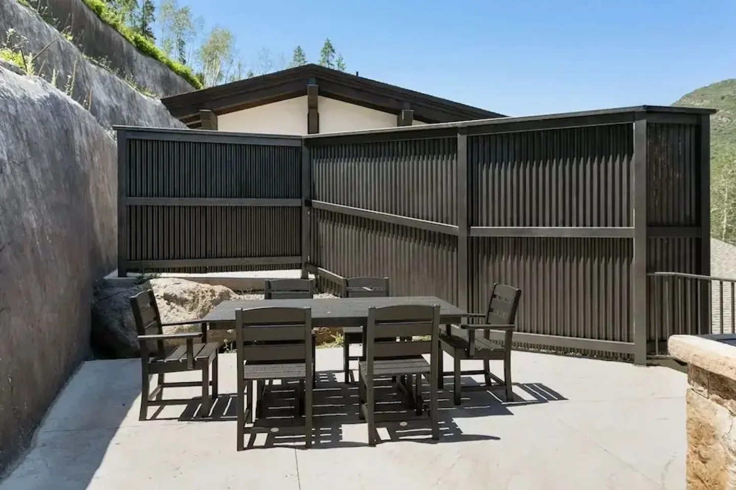 Outdoor patio with a dark dining table and chairs against a slatted privacy fence and rock wall.
